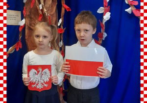 Agatka i Franek w odświętnym ubraniu trzymają w rączkach godło i flagę Polski, w tle dekoracja okolicznościowa